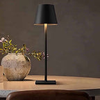 Black rechargeable table lamp on a wooden surface with a vase and flowers in the background 
